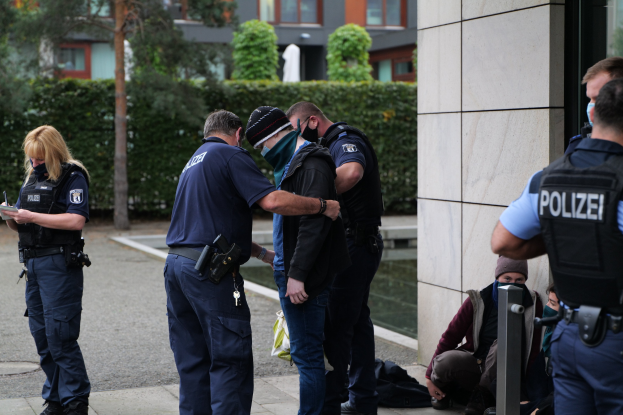 Deutsche Polizeibeamte vor einem Gebäude mit einer Frau, die ein Buch und einen Stift hält, während andere sitzen und stehen, mit Bäumen und Gebäuden im Hintergrund.