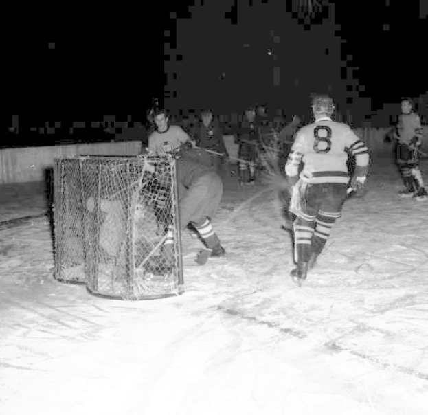 Schwarzes und weißes Foto von Männern, die Hockey auf einem Eisstadion spielen, mit einem Netz im Vordergrund und einer Wand im Hintergrund.
