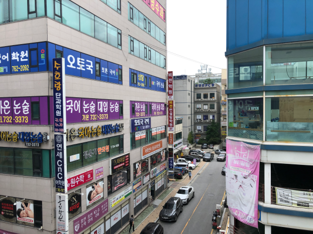Eine belebte Stadtstraße mit hohen Gebäuden, fahrenden Fahrzeugen, Fußgängern auf dem Gehweg, Werbetafeln, Bannern, Bäumen und einem klaren blauen Himmel.
